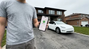 A homeowner in a driveway checking an insurance app on their smartphone with their house and car in the background.